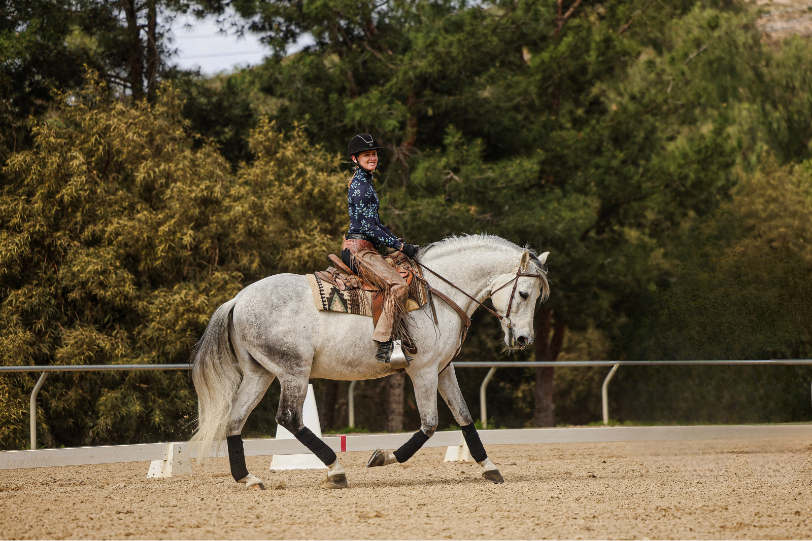 A person wearing a cowboy hat and western attire rides a light gray horse at a graceful canter in an outdoor arena, surrounded by trees and a fence, as if ready to Open Page on a new adventure.