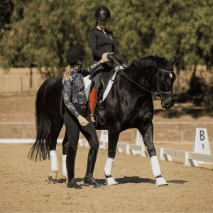 Two women in riding clothes and helmets work with a black horse in an outdoor arena. One woman is mounted, focusing on her rider position, while the other stands beside the horse, offering guidance. White markers and trees are visible in the background.