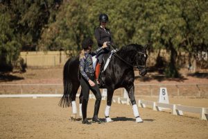 A woman on horseback practices dressage in an outdoor arena, focusing on her Rider Position Blueprint as another woman, walking beside the horse, gives instruction. Both wear riding helmets and black attire. Trees and a fence are in the background.