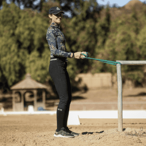 A woman in athletic wear demonstrates a Rider Position as she stretches a green resistance band attached to a metal rail outdoors on a sandy surface, with trees and a gazebo in the background.