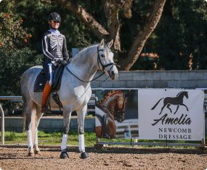A person in a skeleton costume demonstrates excellent rider position while riding a light gray horse near an Amelia Newcomb Dressage sign with a horse logo and photo. Trees and a fence complete the backdrop, making it a great blueprint for spooky equestrian fun.