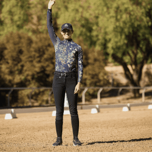 A person wearing a floral long-sleeve shirt, black pants, and a cap stands on a sandy area outdoors, smiling and raising their right arm in a Rider Position Blueprint pose. Trees and a fence are visible in the background.