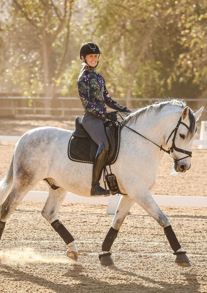 Olivia, in a helmet and patterned top, smiles while riding a white horse in an outdoor sandy arena, surrounded by trees and sunlight—just like the joy of learning at a Free Training or Scale Webinar.