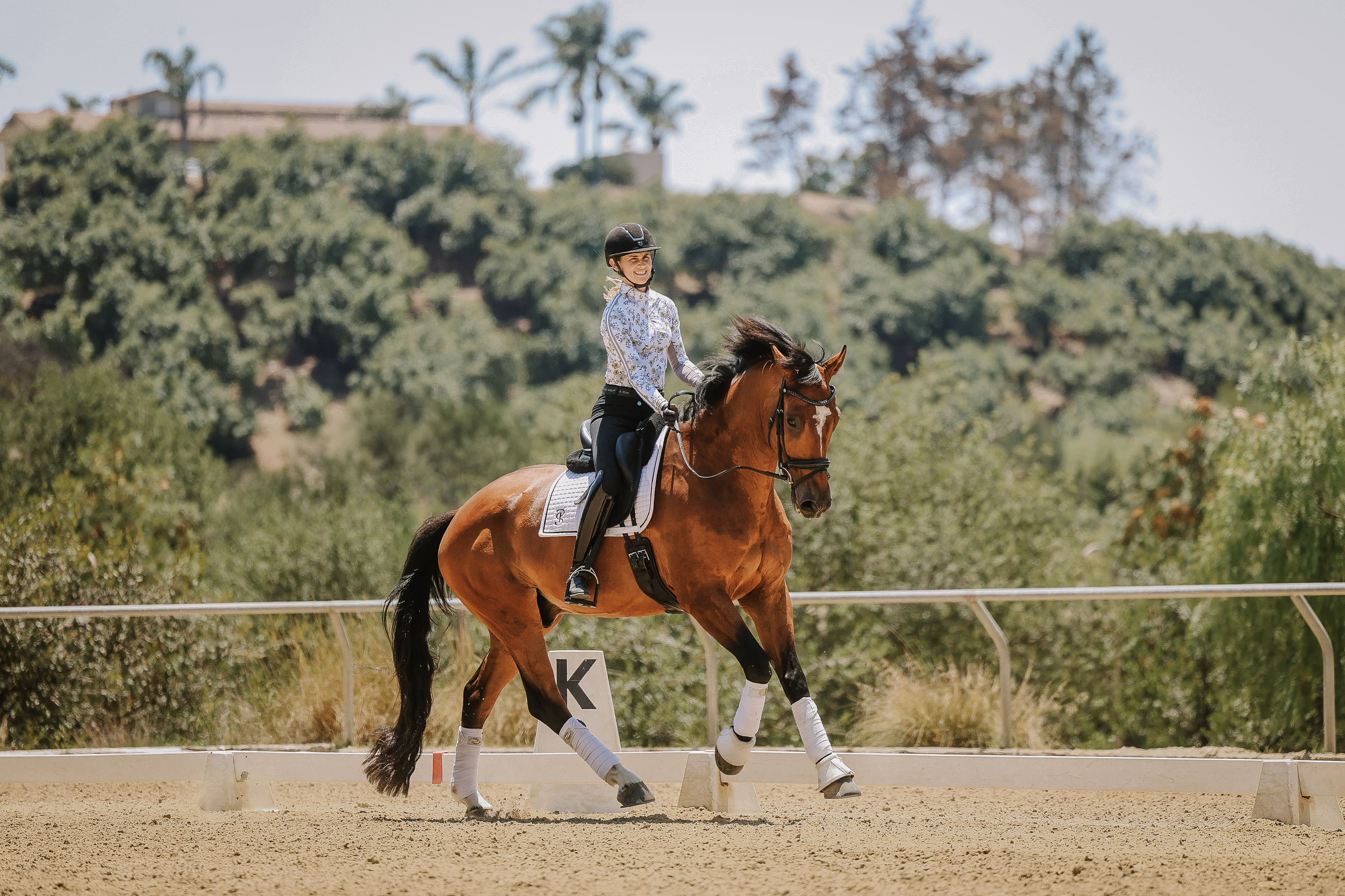 A person in a helmet, blouse, and riding boots practices Dressage Training on a brown horse with white leg wraps in an outdoor arena. Trees and greenery surround the scene beneath a clear sky.