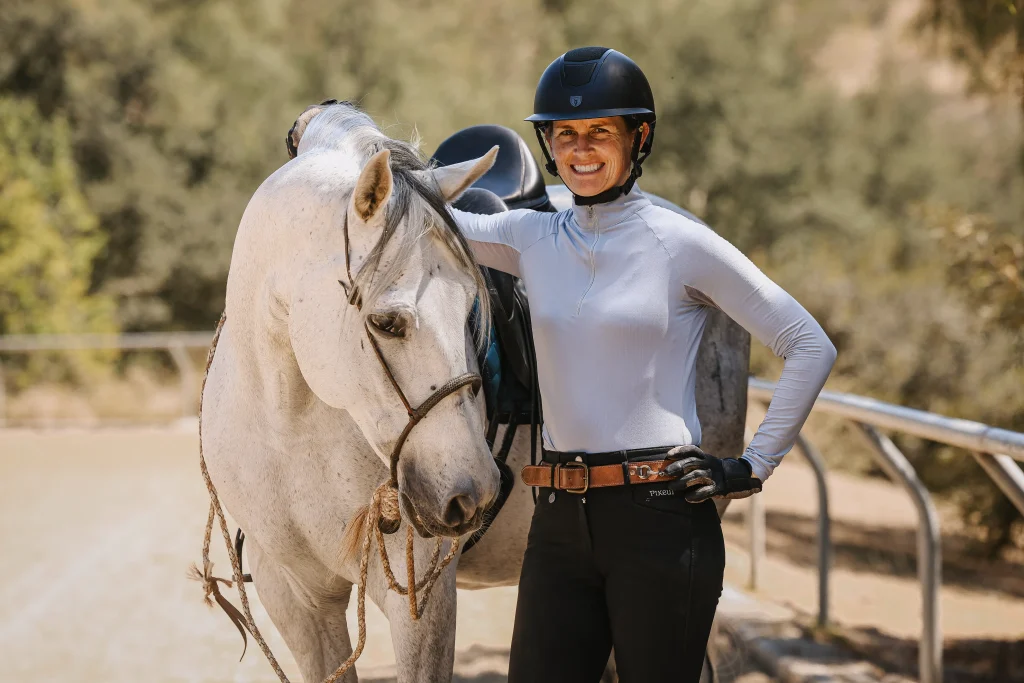 A person wearing a helmet and equestrian gear stands outdoors next to a white horse, smiling with one arm resting on the horse's back. With $297 entry, the 2025 Open is just 30 Days to Round—trees and a metal fence frame the scene.