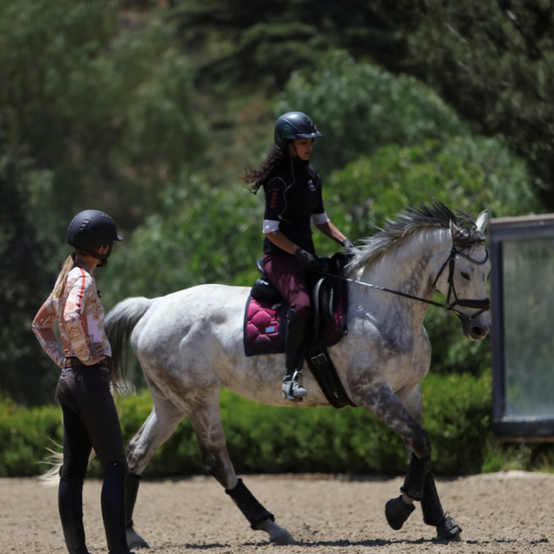 A person rides a gray horse in an outdoor arena, wearing a helmet and equestrian gear, while another person, also in a helmet, stands nearby watching and providing instruction. Trees and greenery are in the background.