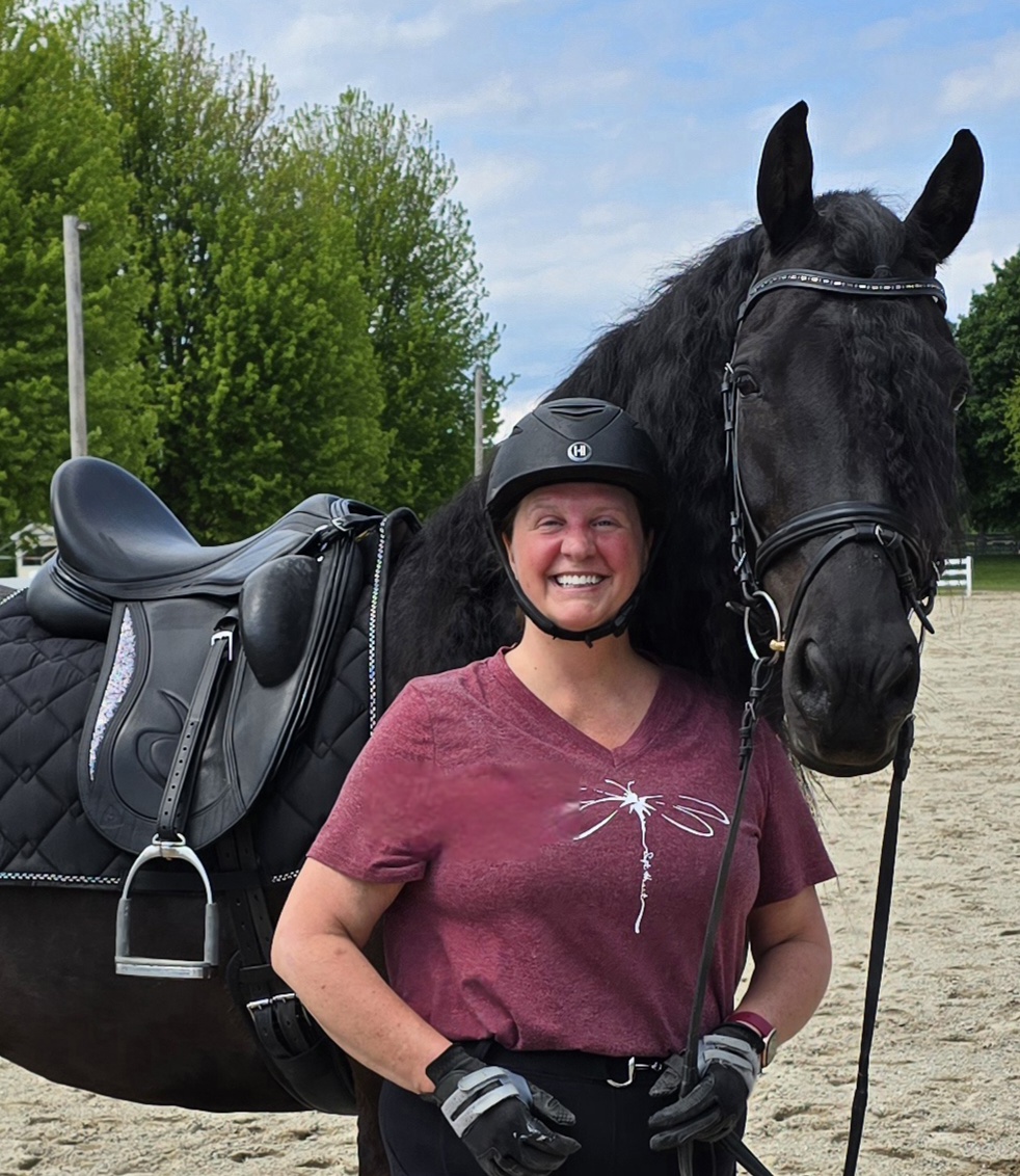 Strides member Kris, smiling in a maroon shirt and black riding helmet, stands beside a black horse with a braided mane on a sandy riding arena, framed by trees in the background.