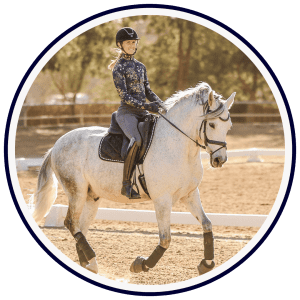Amelia Newcomb, in a helmet and floral jacket, schools a grey horse at trot in a sunny outdoor arena bordered by lush trees.