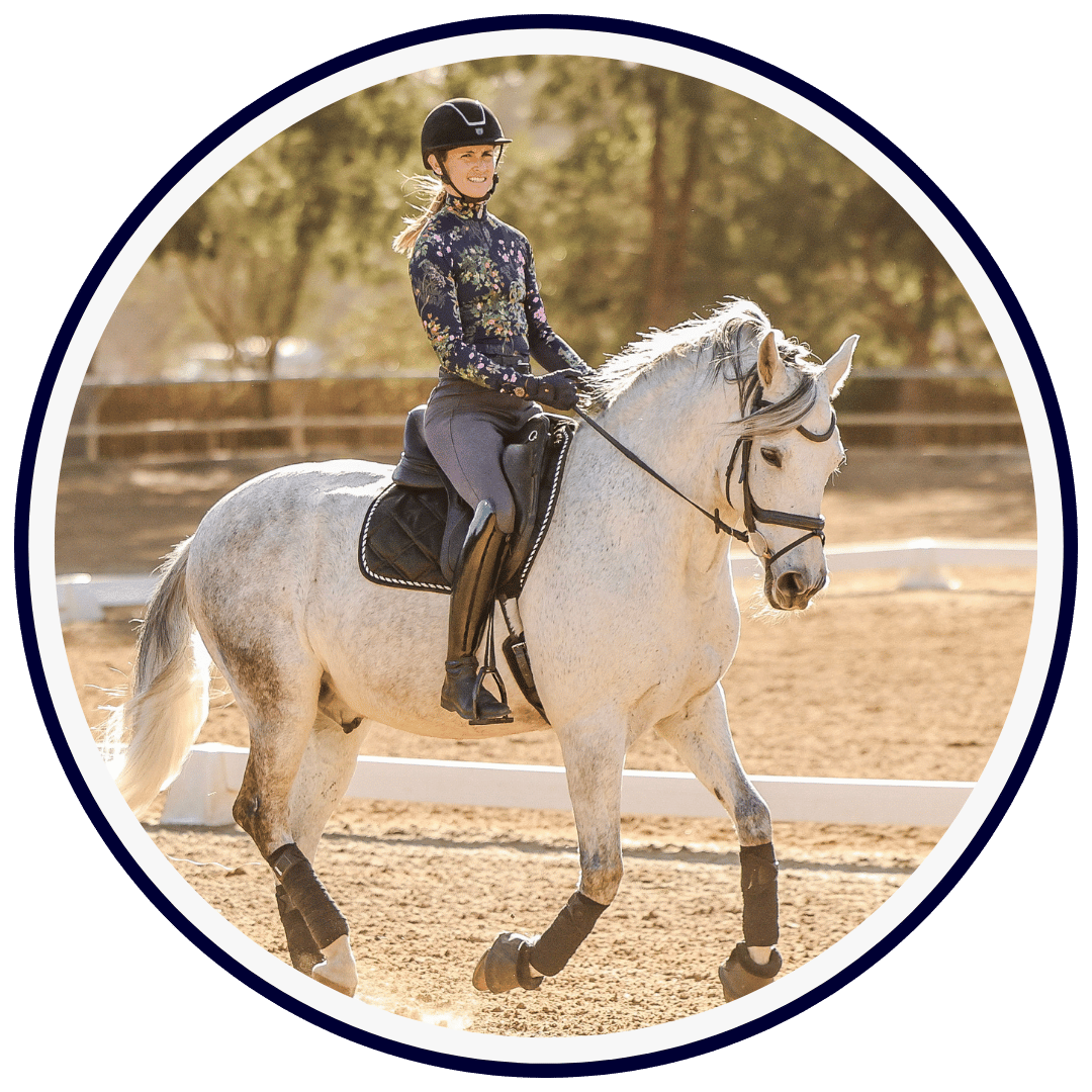 3 Amelia Newcomb, in a helmet and floral jacket, schools a grey horse at trot in a sunny outdoor arena bordered by lush trees.