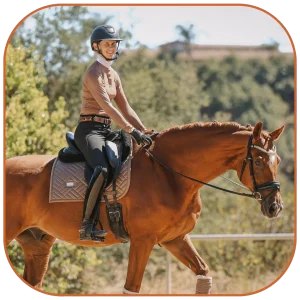 A person wearing a helmet, tan top, and black riding pants is smiling while riding a brown horse outdoors on a sunny day, enjoying contact and connection with nature amid trees and greenery in the background.