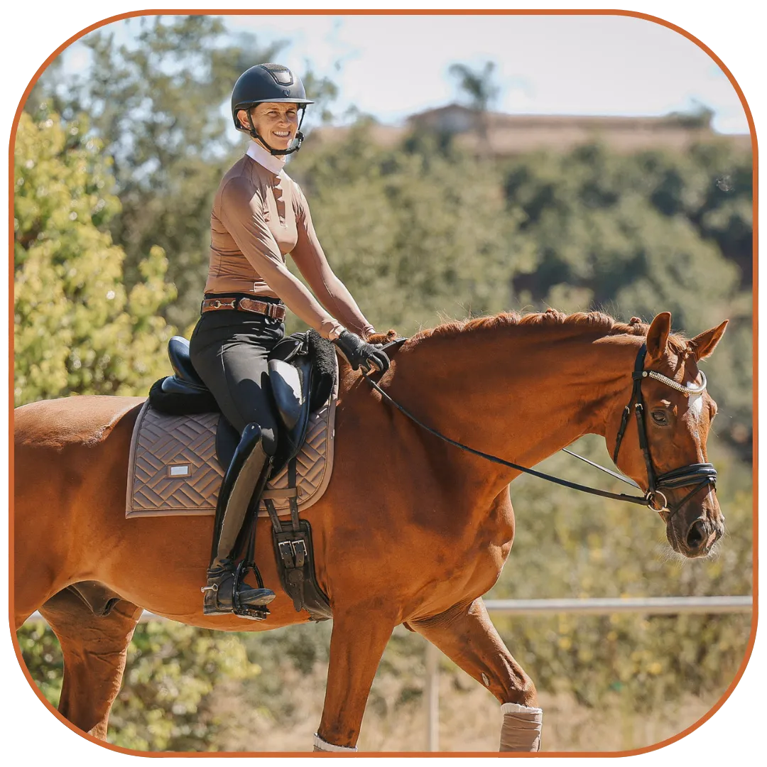 A person wearing a helmet, tan top, and black riding pants is smiling while riding a brown horse outdoors on a sunny day, enjoying contact and connection with nature amid trees and greenery in the background.