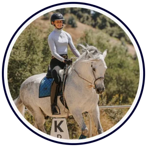 A person wearing a helmet and riding gear is riding a white horse with a blue saddle pad outdoors, passing by a K sign on a sunny day with trees in the background—perfect for illustrating our Free Worksheet on equestrian basics.