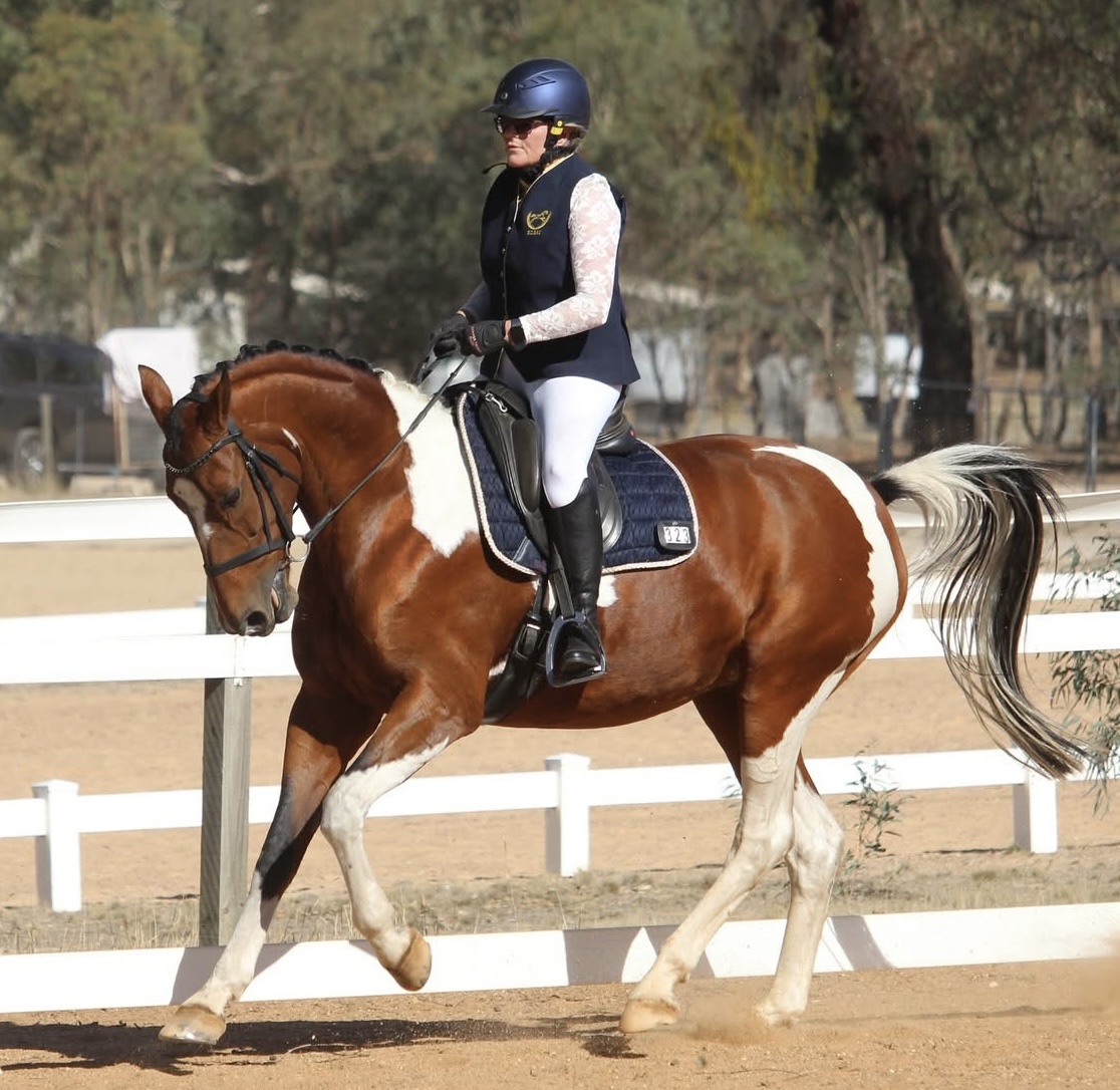 A Strides member named Deidre, wearing a helmet, sunglasses, and riding attire, is riding a brown and white horse in an outdoor equestrian arena with white fences and trees in the background.