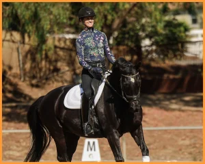 A smiling equestrian wearing a floral long-sleeve shirt, helmet, and gloves practices Free Rider Position Training while riding a black horse with a white saddle pad in an outdoor arena with trees and a fence in the background.
