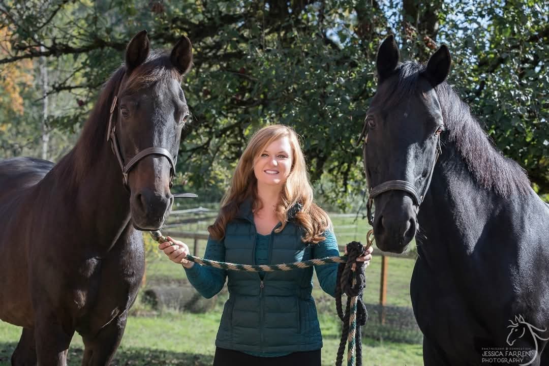 Strides member Shonnessy, a woman with long light brown hair in a teal vest, stands outdoors holding the reins of two large black horses, one on each side, with trees and a fence in the background.