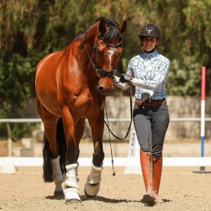 A smiling equestrian, preparing for Round 2025, walks alongside a brown horse with white leg wraps in a sandy outdoor arena, surrounded by greenery and riding equipment—ready for 30 days of training inspired by Oli B.