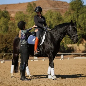 A horseback riding instructor stands beside a rider sitting on a black horse in an outdoor arena, giving guidance. Both wear helmets and riding gear, preparing for the 2025 Open with tips from Oli B's 30 Days to Round. Trees and fencing frame the scene.