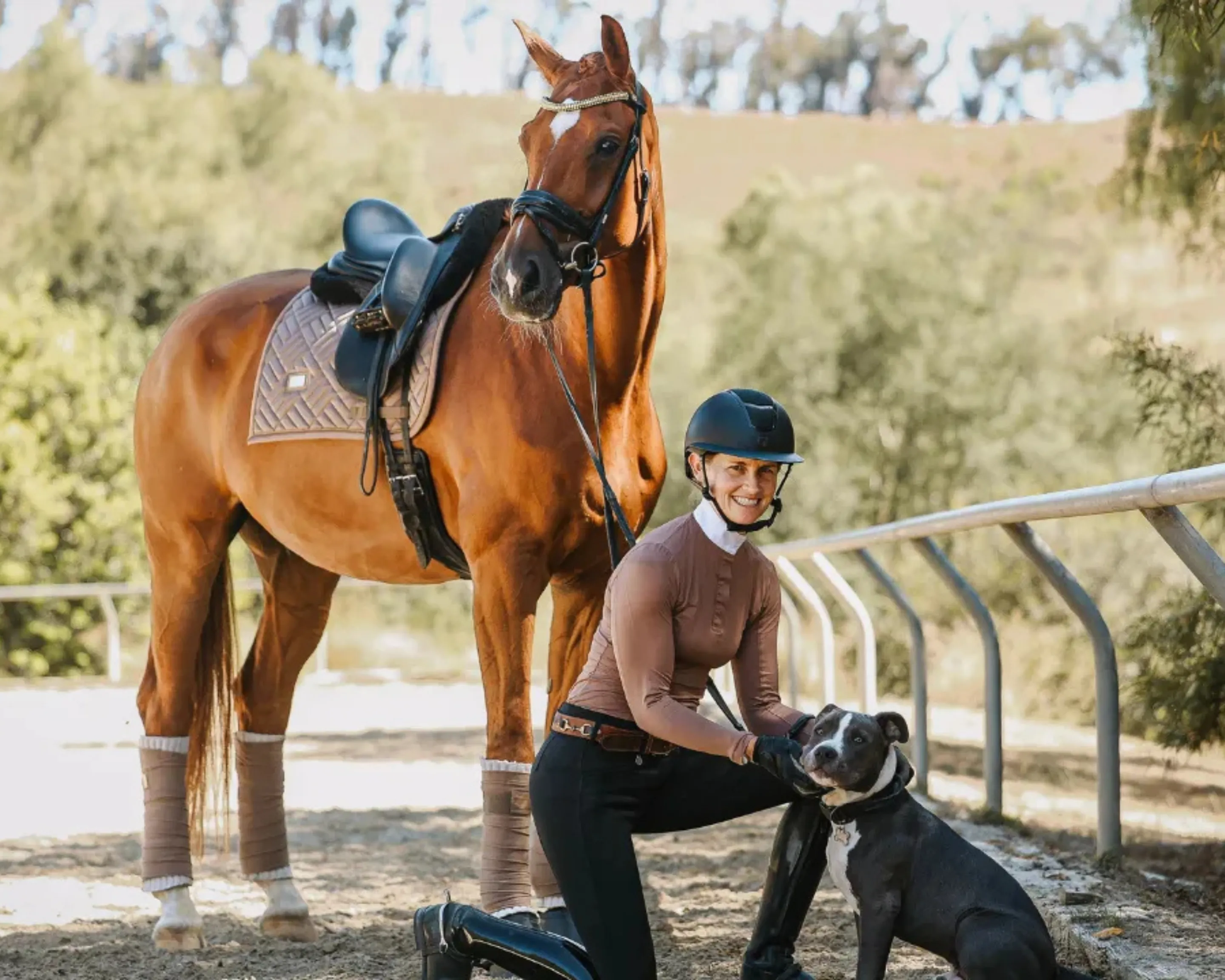 Amelia Newcomb, helmeted, kneels by her black and white dog beside a tacked bay horse at rest outdoors near a paddock fence.
