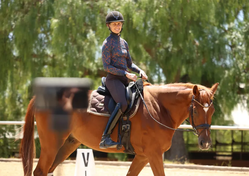 A person in a helmet and patterned shirt rides a brown horse in an outdoor arena, with green trees and a PIVO dressage marker visible nearby; this popular accessory is currently sold out.