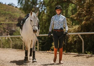 A smiling person in equestrian gear walks beside a white horse along a fenced dirt path outdoors, with trees and hills visible in the background—perfect inspiration for our Free Groundwork Webinar.