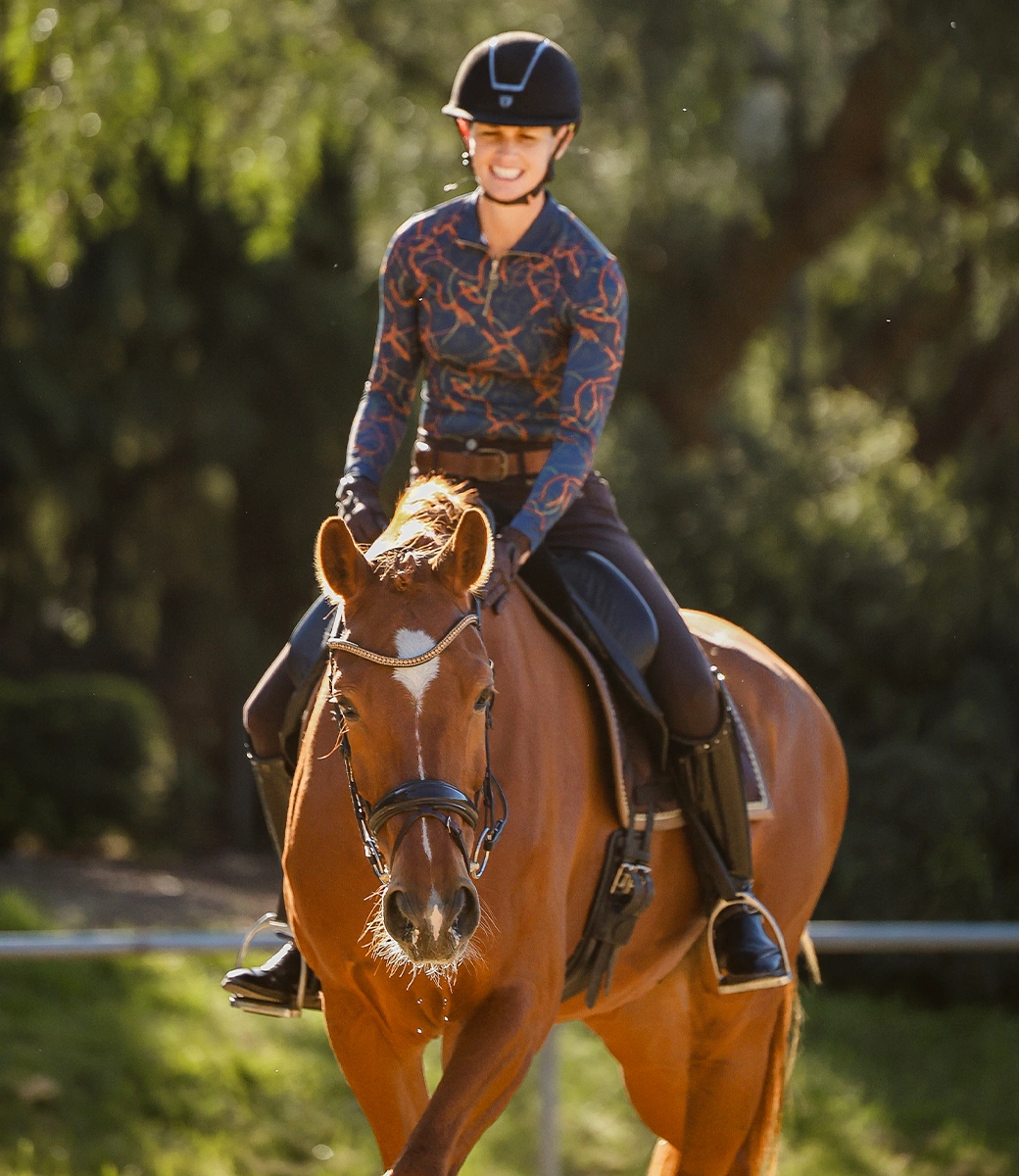 A person wearing a helmet and patterned shirt smiles while riding a brown horse outdoors on a sunny day, with green trees in the blurred background.