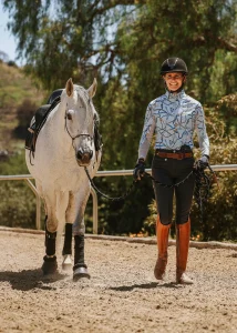 A smiling equestrian walks alongside a white horse on a sandy path, demonstrating Groundwork Training while holding its reins. The rider wears a helmet, patterned shirt, and tall brown boots. Trees and greenery are visible in the background.