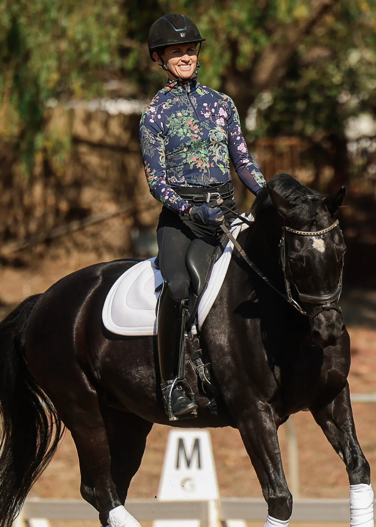 A smiling equestrian in a floral shirt, helmet, and riding gear sits atop a black horse with a white saddle pad, outdoors near a riding arena marker labeled M. Trees and sunlight are in the background.