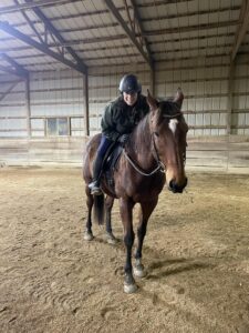 Strides member Wendi, wearing a helmet and dark clothing, is sitting and smiling on a brown horse inside a large indoor riding arena with a dirt floor and wooden walls.