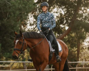 A person in a helmet, patterned jacket, gloves, and riding pants is riding a brown horse in an outdoor area surrounded by trees and sunlight, practicing movements inspired by classic Dressage Terms.