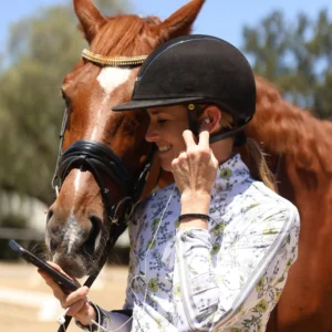 A woman in a riding helmet and floral shirt smiles while holding a phone and adjusting her earbud, standing beside a brown horse with a bridle and decorative browband outdoors during a holiday workshop.