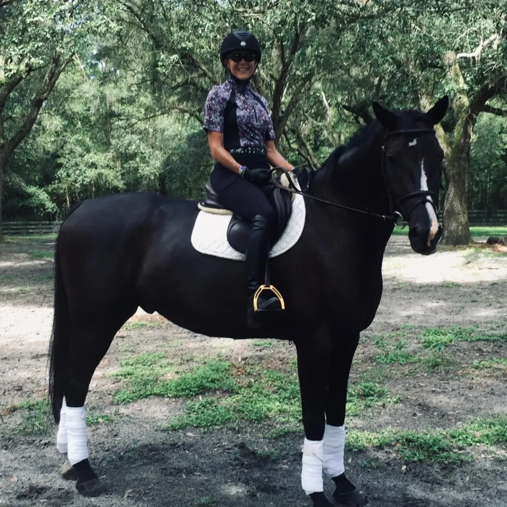 A person wearing a helmet, sunglasses, and riding gear sits on a dark horse with white leg wraps and a white saddle pad in a wooded outdoor area.