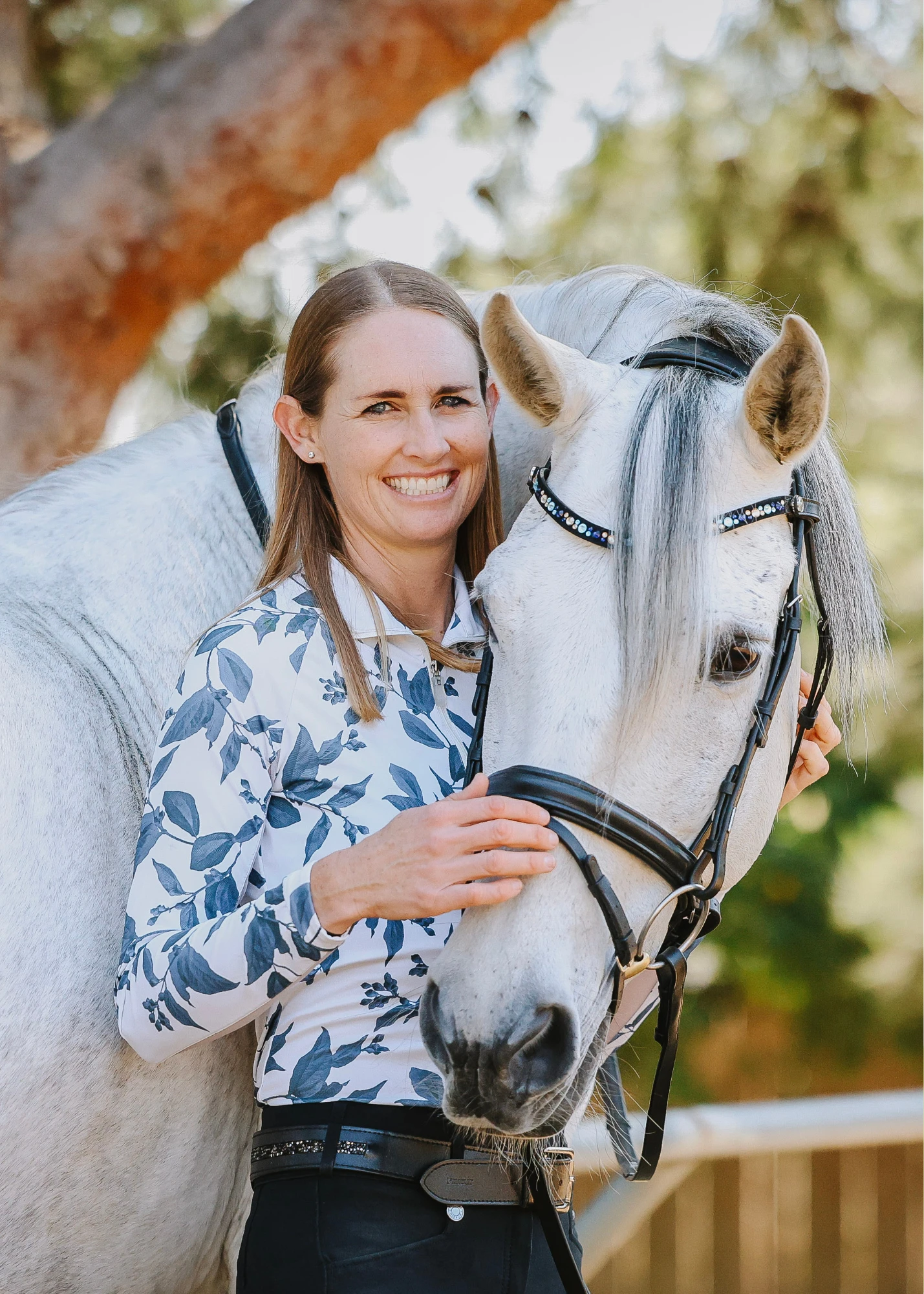 A smiling woman with straight, light brown hair stands beside a white horse, gently holding its face. Outdoors amid sunlit trees, she wears a floral-patterned shirt—capturing the harmony at the heart of Dressage Foundations.
