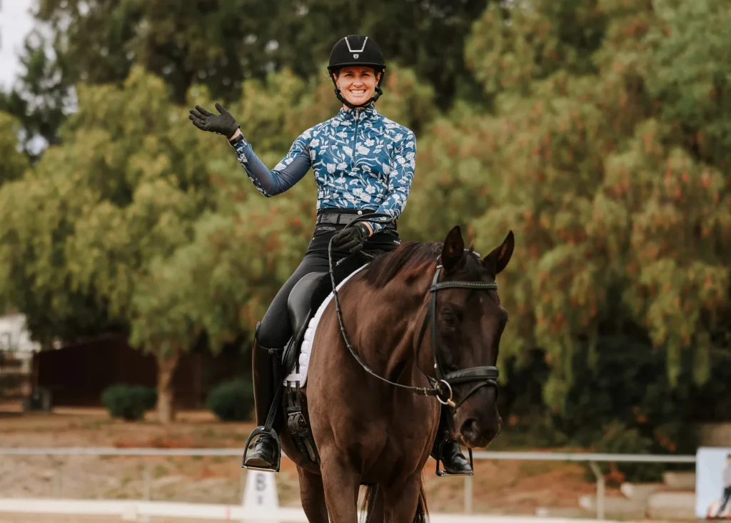 A person wearing a helmet and patterned jacket smiles and waves while sitting on a dark horse outdoors, with green trees and a fenced area in the background—perfect for highlighting Dressage Foundations on your Sale Page.