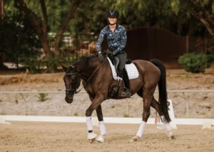 A smiling equestrian wearing a helmet, patterned shirt, and gloves rides a dark horse with white leg wraps and a white saddle pad in an outdoor arena—perfecting her Dressage Foundations amid trees and a fence in the background.