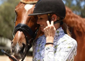 A woman wearing a helmet and floral shirt smiles while adjusting her earbud, holding a phone. During her holiday, a brown horse with a bridle nuzzles close to her, their faces touching affectionately.