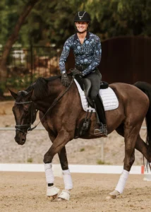 A smiling person in a patterned blue shirt, black helmet, and gloves rides a dark brown horse with white leg wraps and a white saddle pad in an outdoor arena—a perfect moment capturing Dressage Foundations for your Sale Page. Trees and a fence are visible behind.