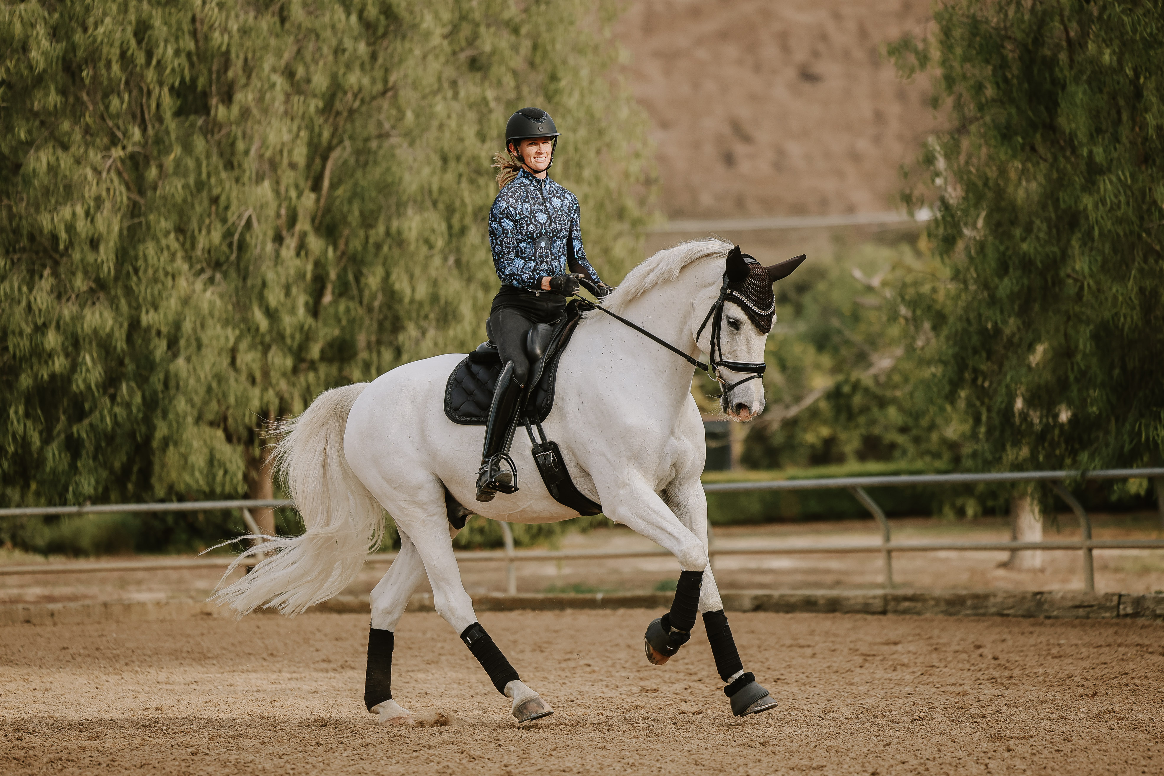 A person wearing a helmet and patterned shirt rides a white horse with black tack and leg wraps in an outdoor arena, surrounded by trees and a fence, during dressage lessons.