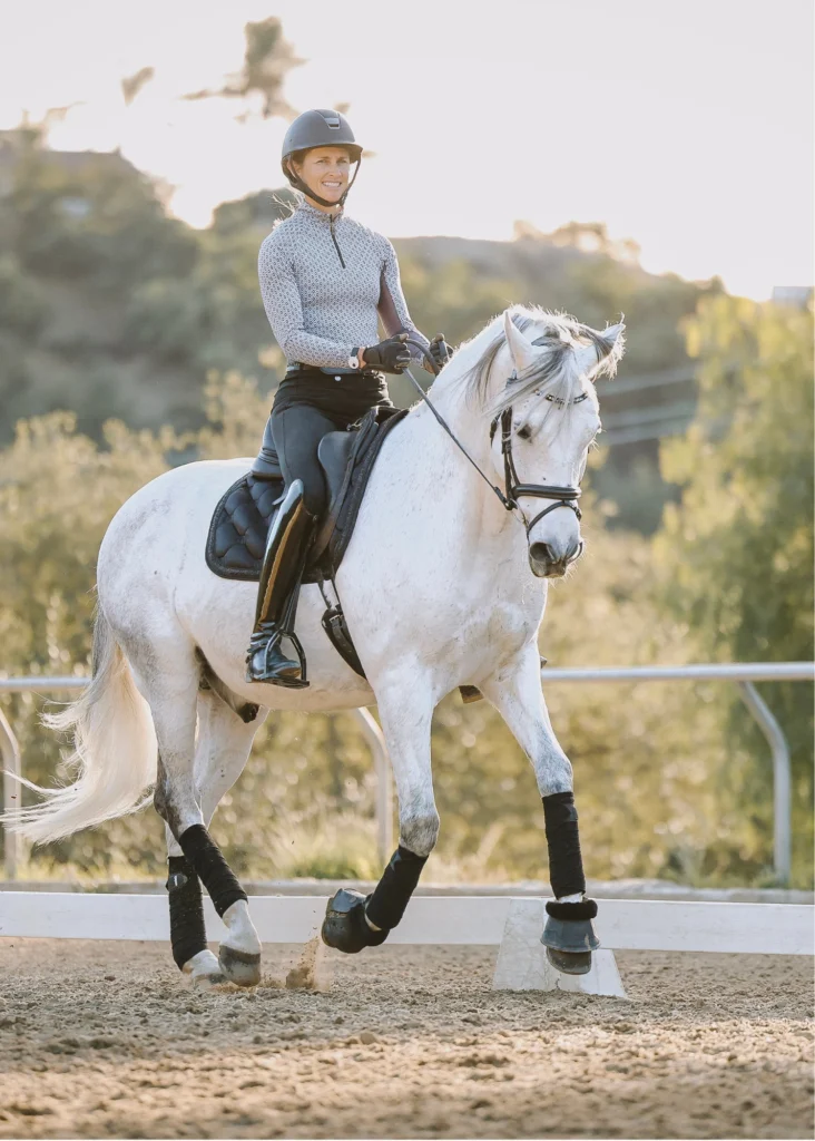 A person in riding gear and a helmet is riding a white horse in an outdoor arena, demonstrating the Training Scale. The horse is trotting with focus, while trees and a fence form the backdrop—a scene fit for a Scale Masterclass.
