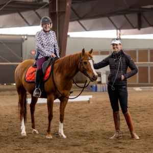 A smiling woman wearing a helmet sits on a chestnut horse with an orange saddle pad, while another in riding gear stands beside them in an indoor arena—celebrating Dressage Foundations starting July 2026. Early Bird spots available!.
