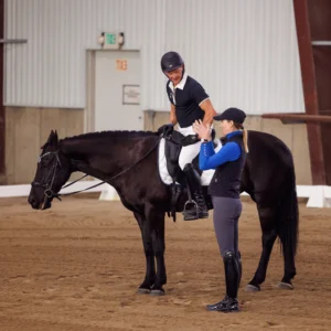 A rider wearing a helmet sits on a black horse inside an indoor arena, smiling at a standing instructor who is clapping and giving feedback during the Dressage Foundations session. The arena has sandy ground and a metal wall in the background.