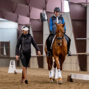 A person on horseback practices Dressage Foundations in an indoor arena while another offers guidance beside them. The rider wears a helmet and vest; the walker wears a hat, jacket, and tall boots. A sign marked E is visible.