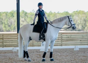 A person wearing a helmet and patterned shirt sits smiling on a white horse with black tack in an outdoor riding arena, ready to start their Dressage Foundations journey amid wooden fencing and trees—perfect for early bird riders this July 2026.