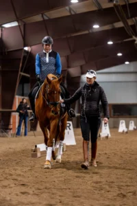 A rider on a chestnut horse practices with confidence in an indoor arena, while a trainer walks beside them guiding and supporting. Both wear riding gear. Dressage markers and sandy footing are visible.