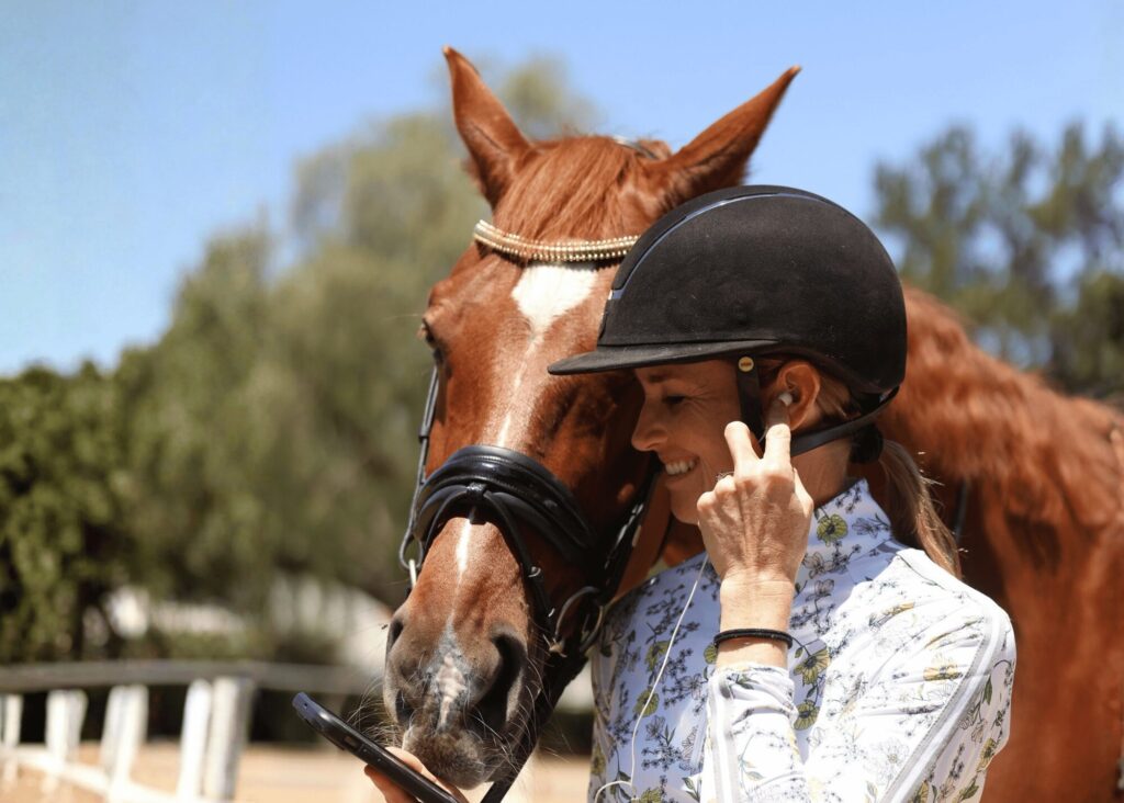 A woman wearing a riding helmet smiles while standing next to a brown horse. The horse nuzzles her as she holds a smartphone—perhaps exploring expert training or Masterclasses—with a fence and trees in the background on a sunny day.