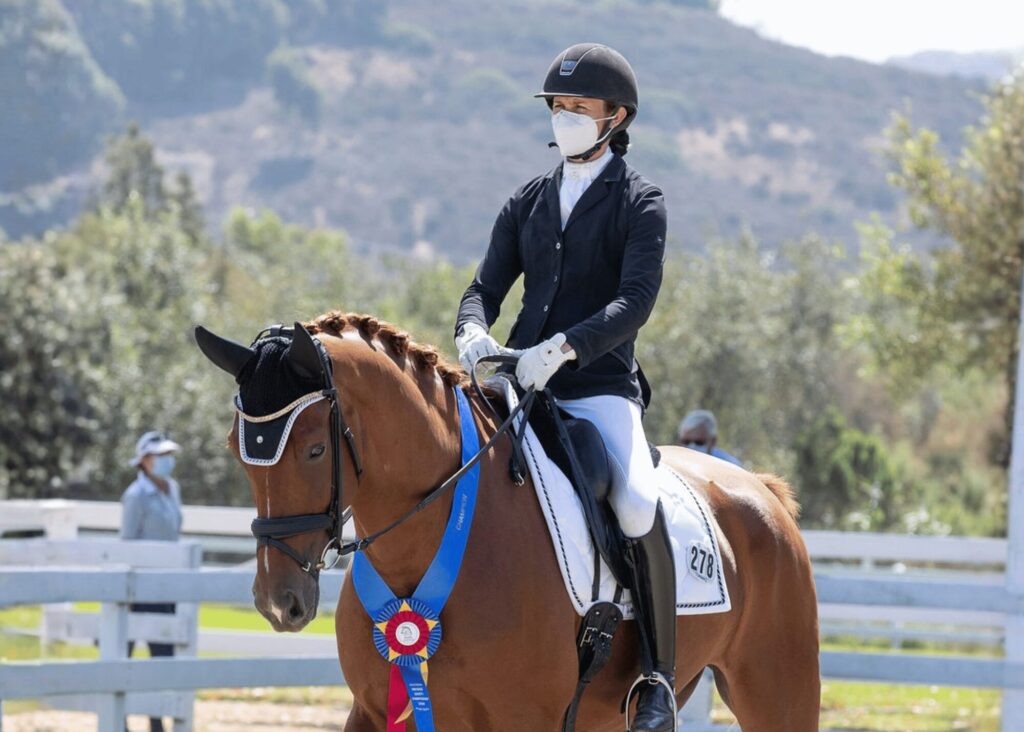 A masked equestrian in formal riding attire sits atop a brown horse wearing a winner’s blue ribbon during a competition, with a sunny outdoor arena and hills in the background—poised as if ready for expert instruction or Masterclasses.
