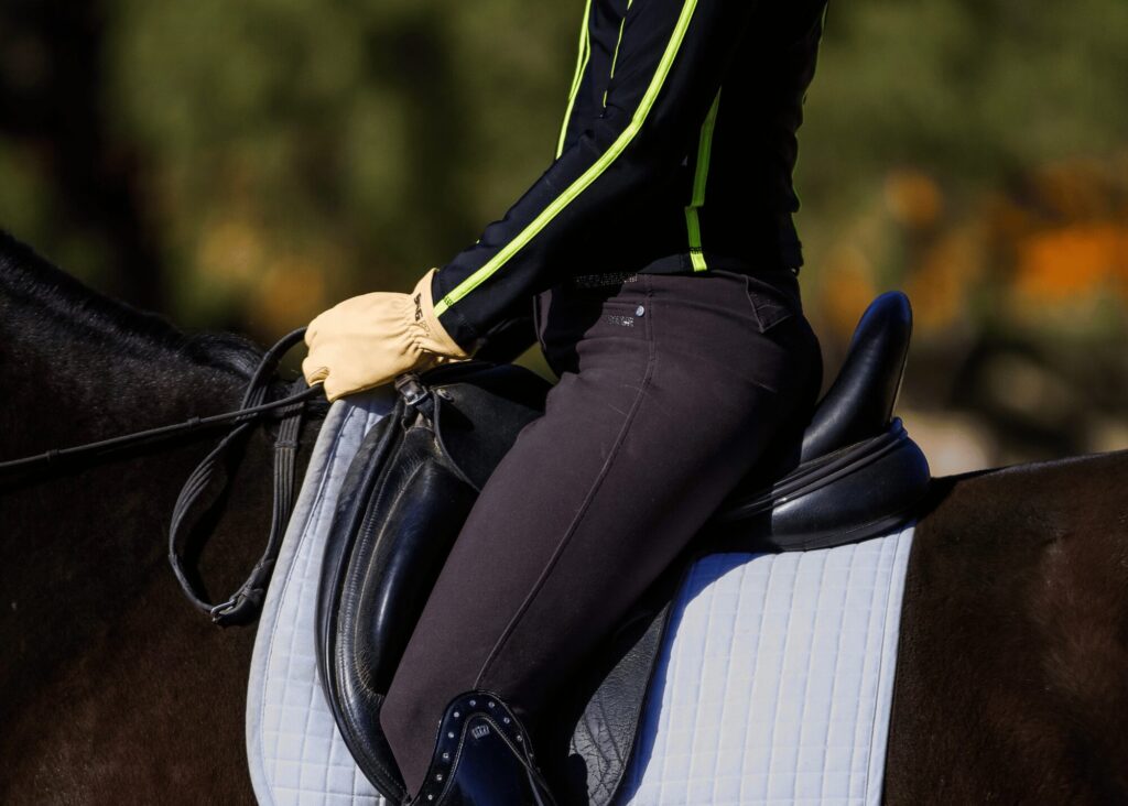 A close-up of a person wearing gloves, dark pants, and a black jacket with neon stripes riding a horse—perfect form for Masterclasses. The focus is on the rider’s torso and hands holding the reins on a padded saddle.