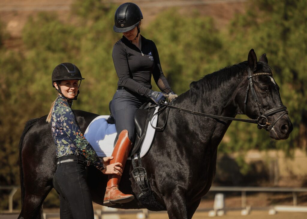 Two women in riding helmets are with a black horse outdoors. One is mounted while the other, possibly an instructor from riding masterclasses, stands beside them, holding the rider’s boot and smiling at the camera. Trees and fencing are in the background.