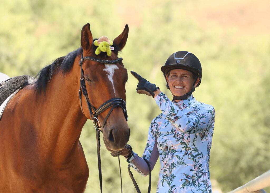 A smiling person in a floral shirt and riding helmet stands beside a brown horse with a white blaze. The horse has a small yellow stuffed toy on its head, and the person is pointing at the toy, as if giving playful masterclasses in horsemanship.