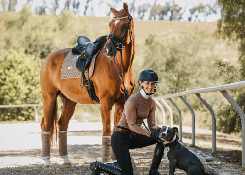 A smiling person in riding gear kneels next to a dog on a sandy path, with a saddled chestnut horse standing beside them—an inviting scene reminiscent of outdoor masterclasses. Trees and greenery are visible in the background.