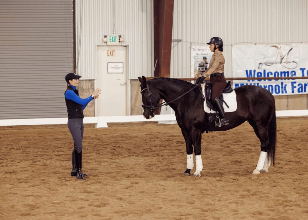 A horse-riding instructor leads a masterclass in an indoor arena, giving instructions to a rider on a black horse. The rider and horse face the instructor, with a Welcome banner visible in the background.